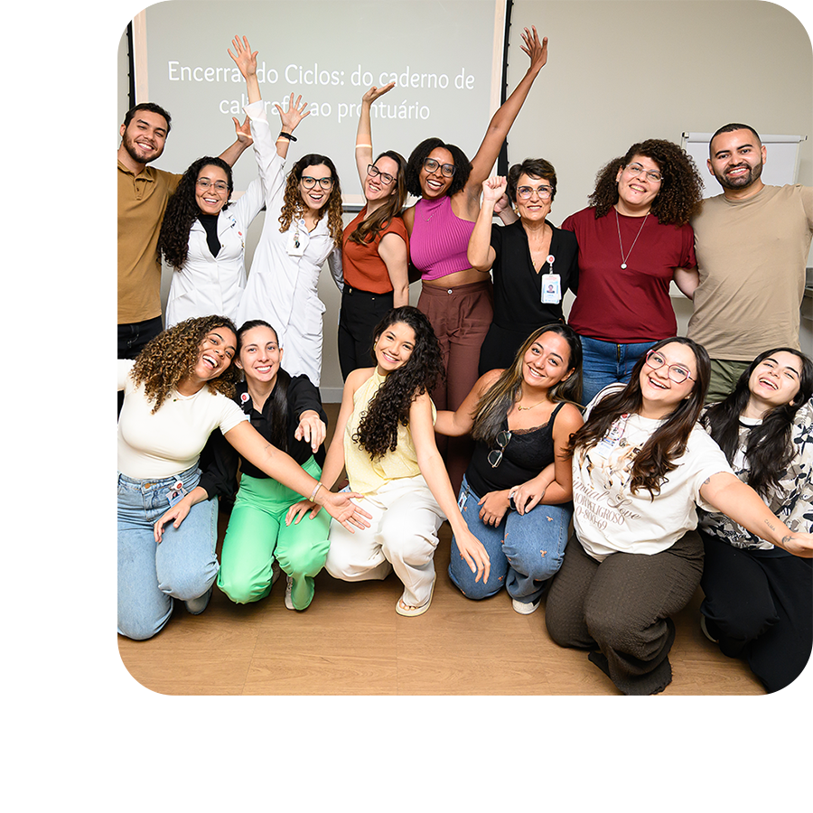 Grupo de pessoas em ambiente institucional posa para foto de forma descontraída, com braços levantados, diante de tela com apresentação ao fundo.